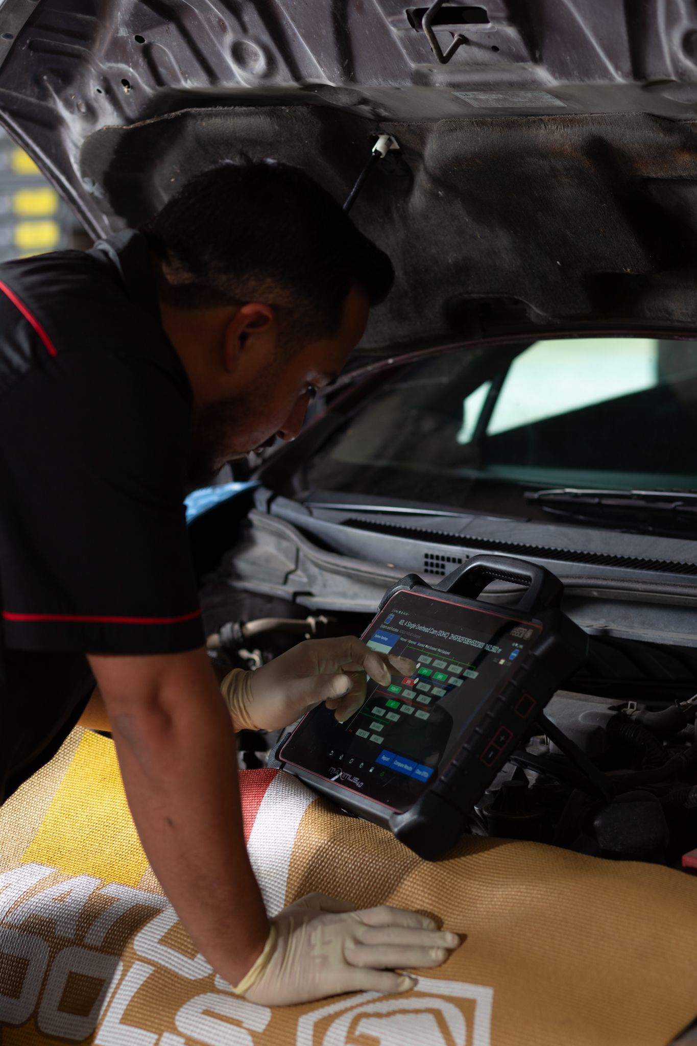 Mechanic using diagnostic tool on car engine, under the hood. Gloves, dark shirt, workshop setting.