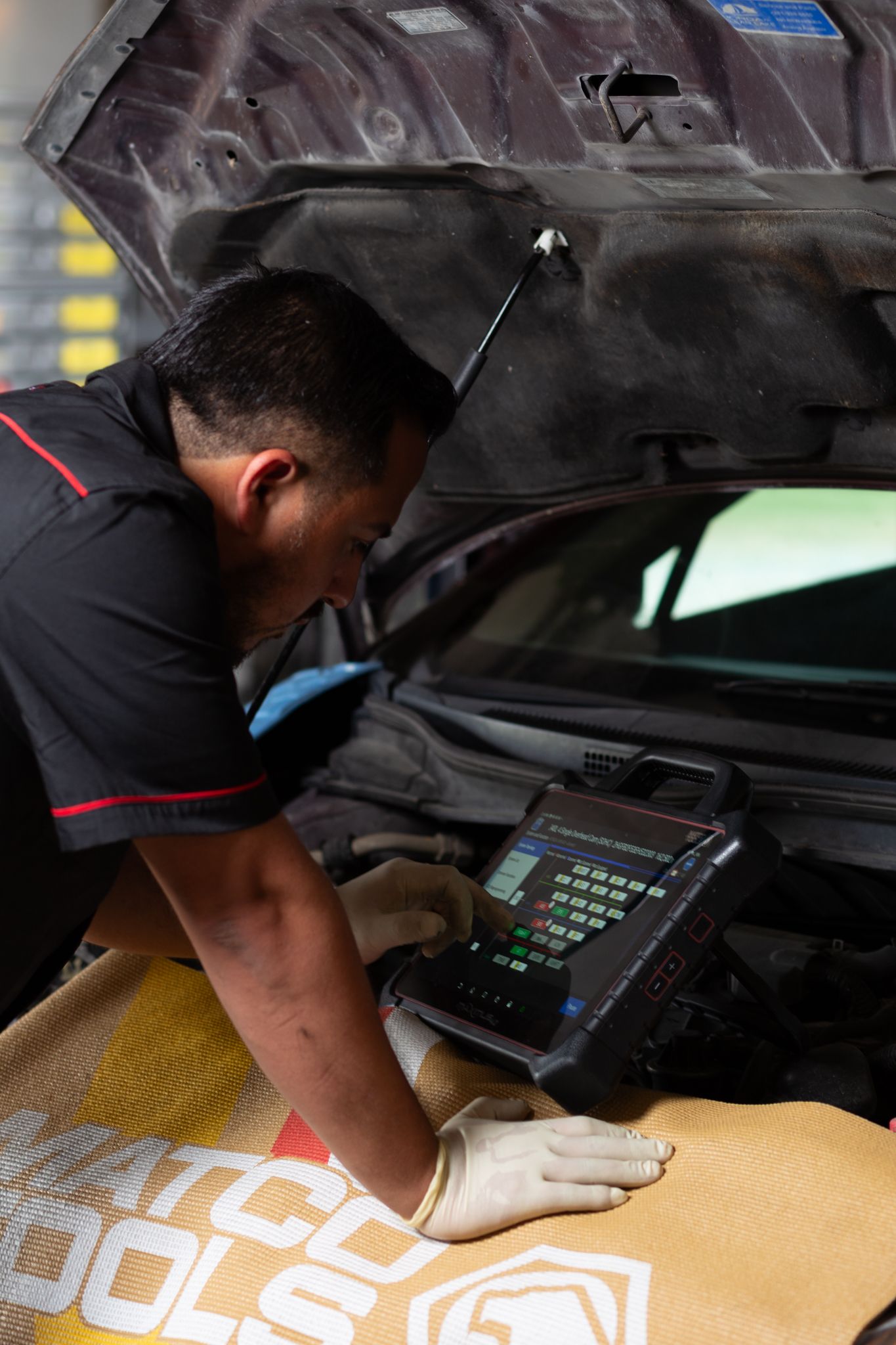 Mechanic using diagnostic tool under car hood. Gloves, dark shirt, in a garage.