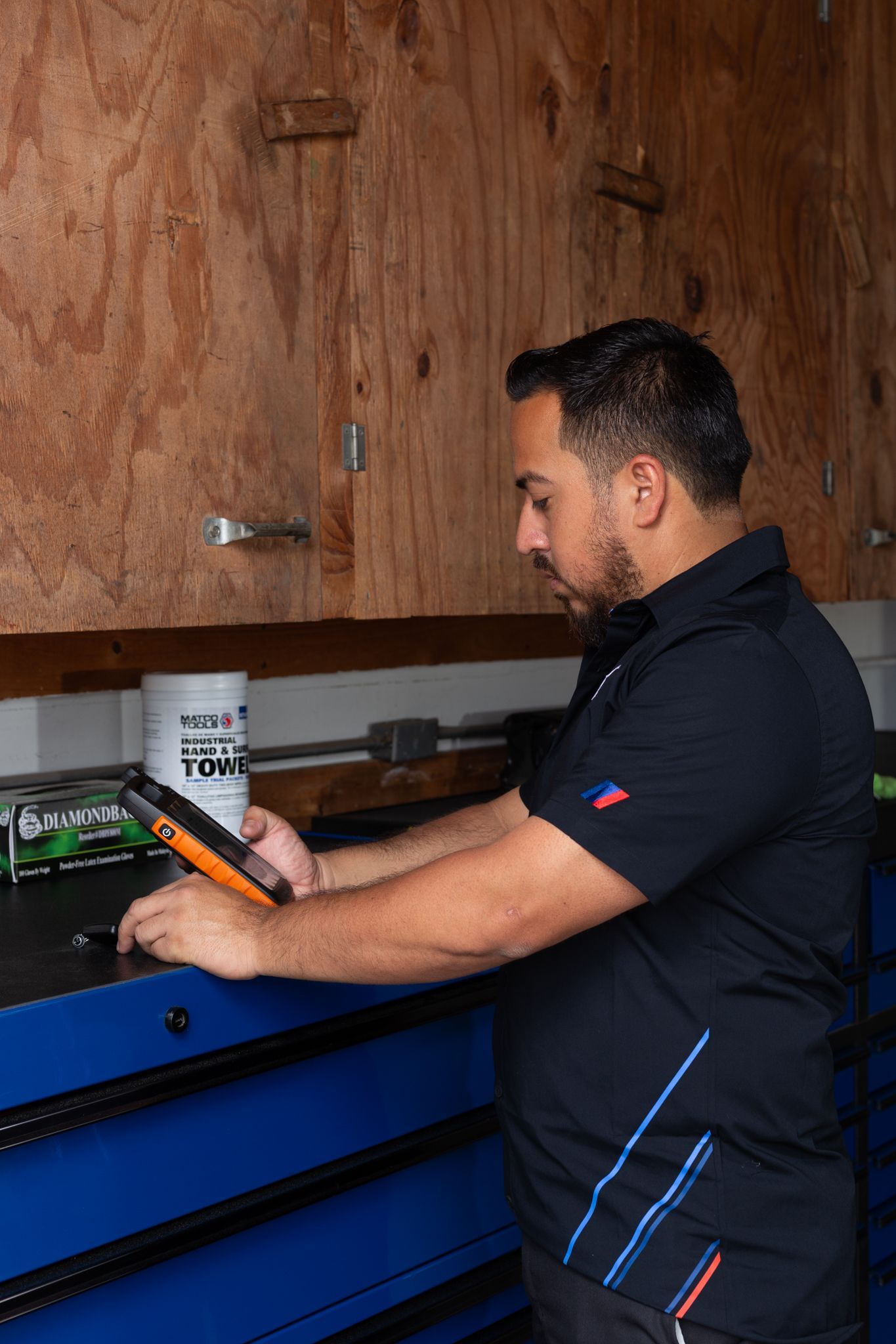 Man in black shirt using a tablet at a workbench in front of a wood-paneled wall.