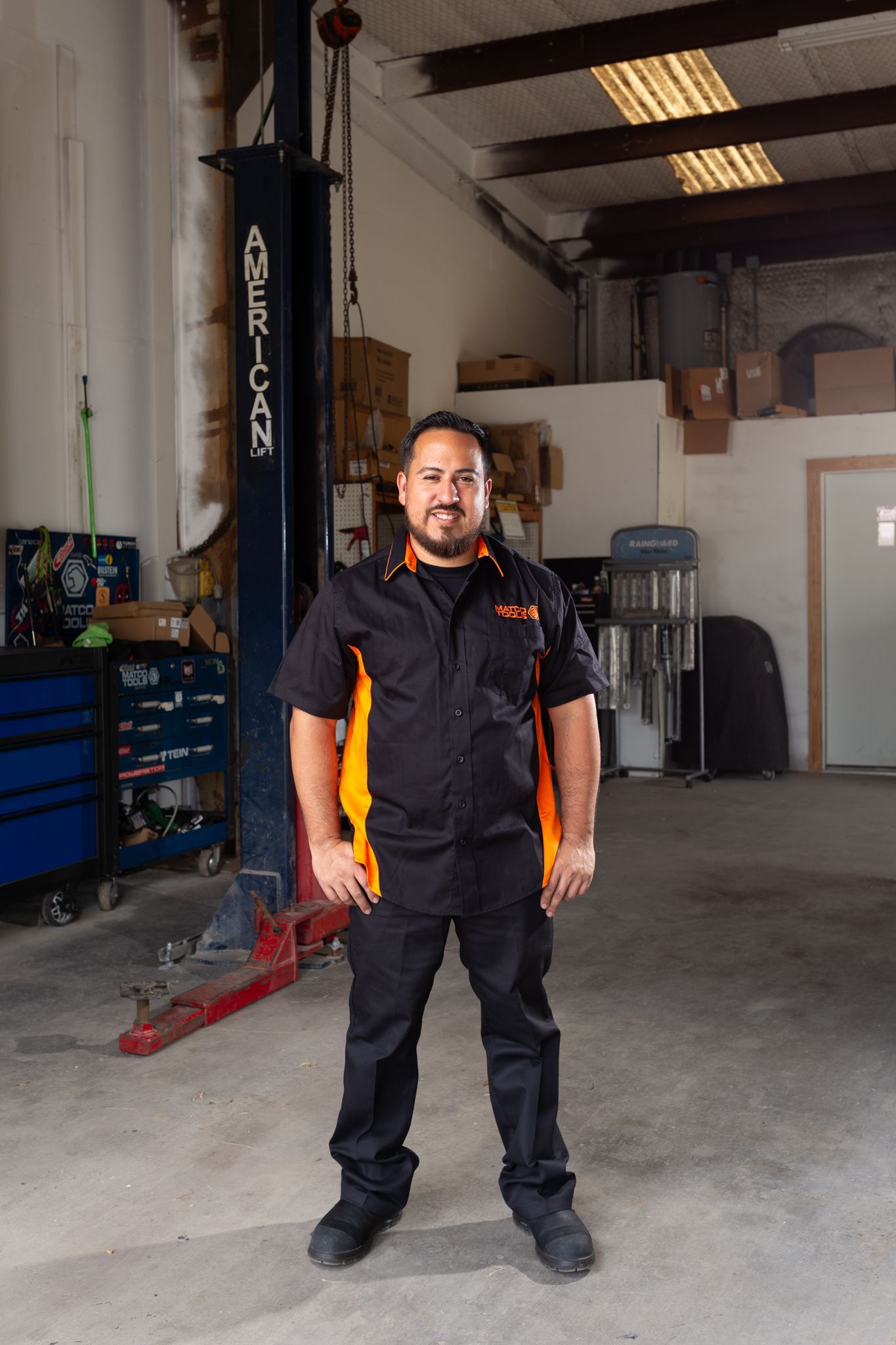 Mechanic in black and orange work uniform stands in a garage; a car lift is behind him.