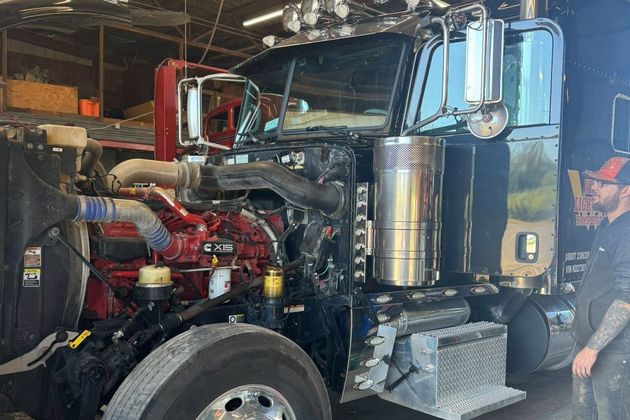Blue semi-truck with hood open in a garage, showing the engine and a mechanic standing beside it.