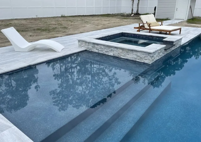 Swimming pool with jacuzzi, deck, and lounge chairs. Blue water, light stone deck, white fence, and greenery.