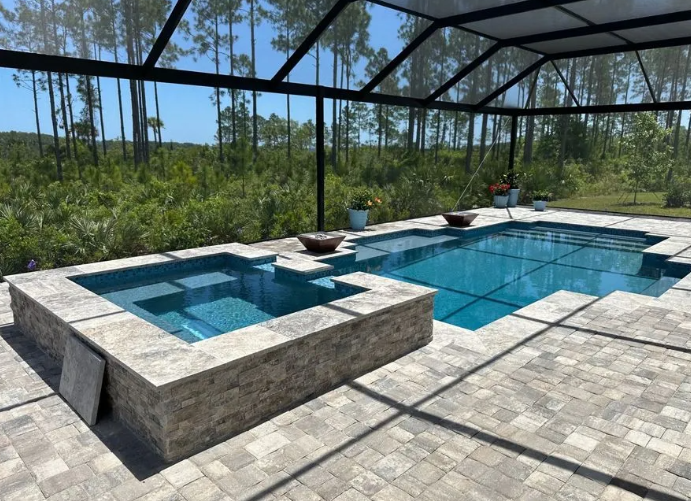 Pool and hot tub with blue water, surrounded by grey stone and screened enclosure. Forest in the background.