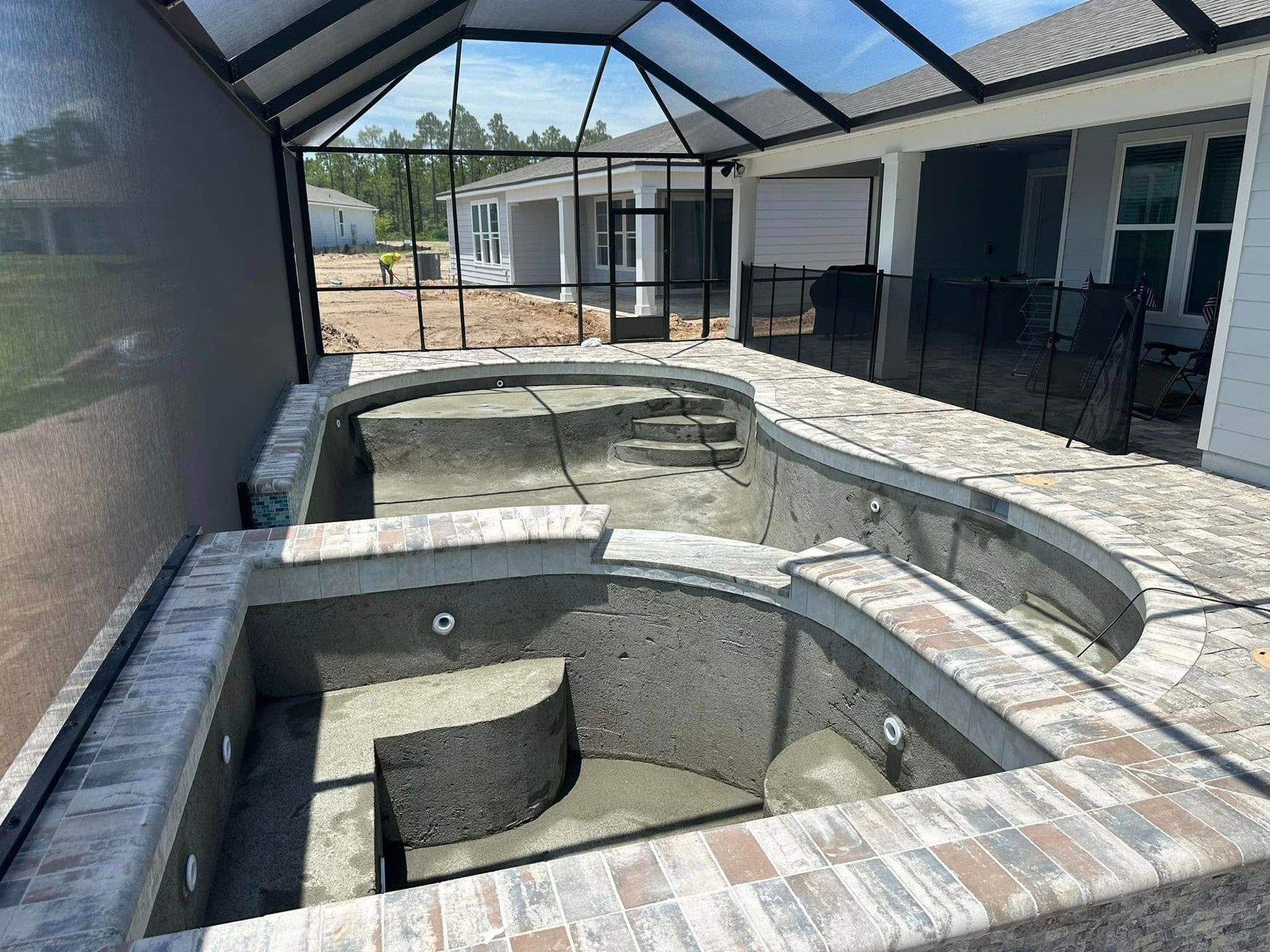 Empty swimming pool and spa under construction, brick coping, screen enclosure, and house in background.