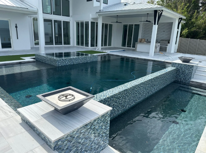 A luxurious backyard pool with dark turquoise tile, a fire pit, and a white house in the background.