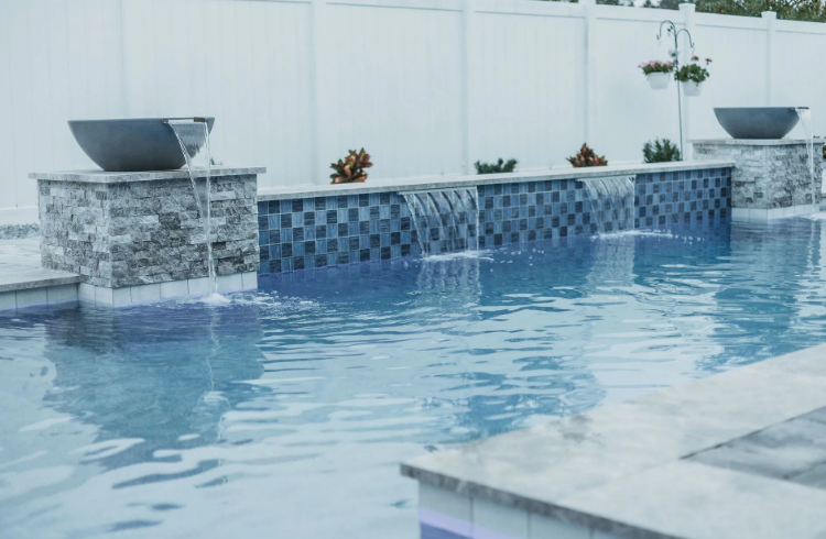 Pool with cascading waterfall feature; stone and tile detail, white fence backdrop.