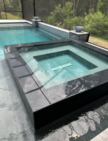 An elevated square spa and adjacent pool with clear water, surrounded by dark tile and greenery.
