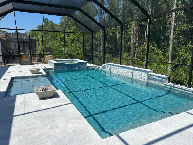 Pool area with screened enclosure, pool, spa, and stone patio. Lush green trees in background.