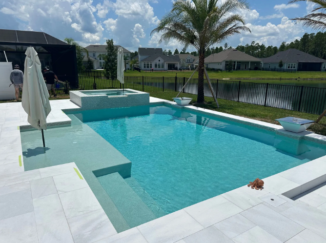 A turquoise pool with a spa. White patio, palm tree, and a black fence.