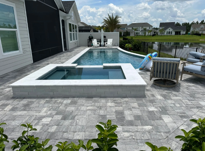 Backyard pool with a spa, paved patio, and lounge furniture. Lush greenery and water views.