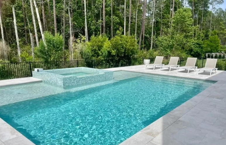 A rectangular pool and jacuzzi with lounge chairs sit on a patio against a backdrop of trees.