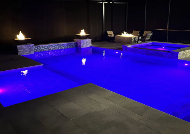 Night view of a luxurious swimming pool with blue lights, fire features, and a hot tub.