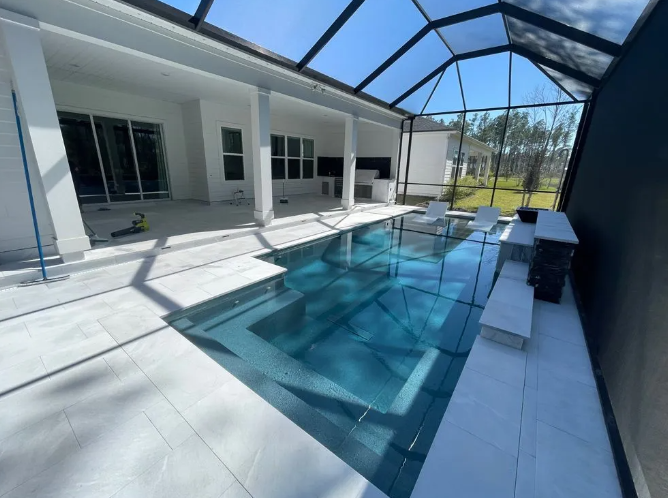 Pool and patio area under a screened enclosure. Blue tiled pool with a built-in bench, white tile deck.