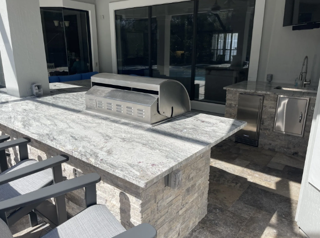 Outdoor kitchen with a stainless steel grill on a granite countertop, with seating and cabinets.