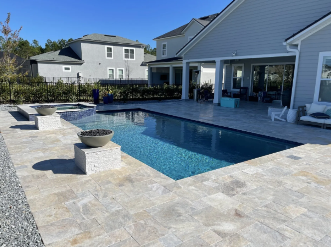 Backyard pool with stone patio, spa, and seating under a covered porch; blue water and gray house.