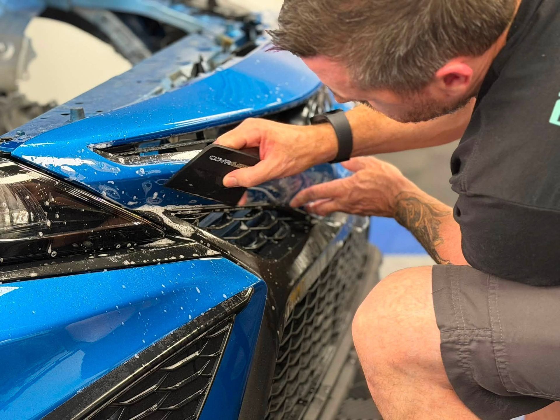 Man repairing a blue car front bumper with a tool in a garage