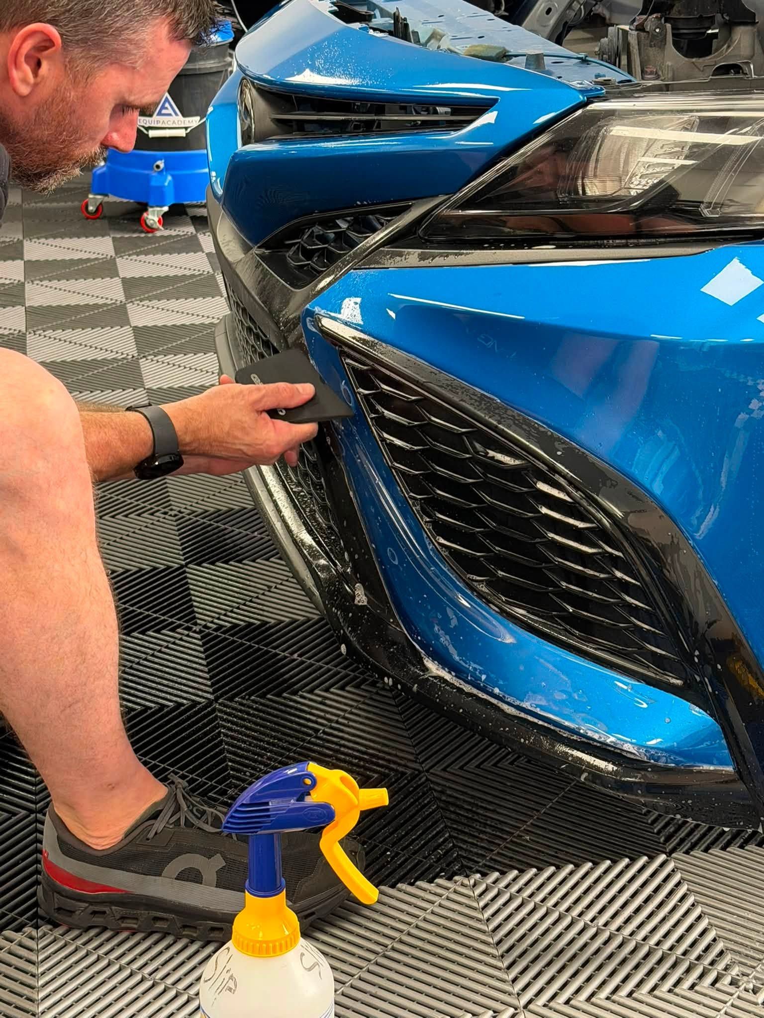 Mechanic inspecting blue car bumper in a workshop, with a spray bottle in the foreground