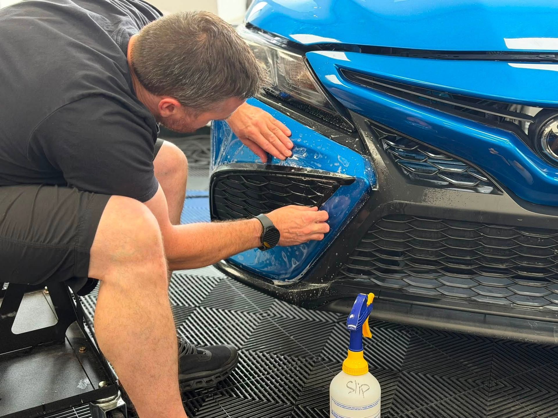 Man applying protective film to the front bumper of a blue sports car in a workshop