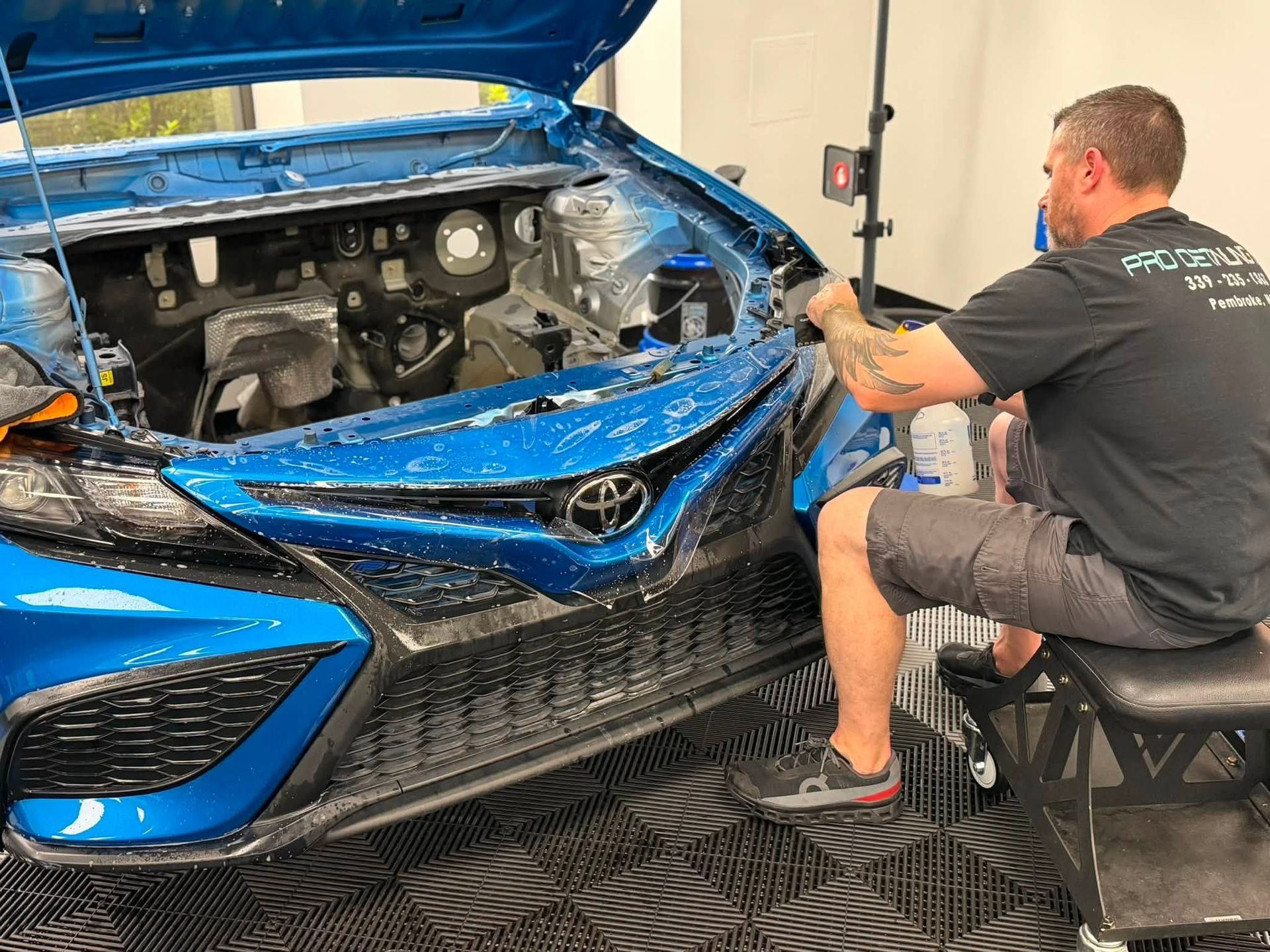 Mechanic repairing the front of a blue car in a shop, with the hood open and bumper removed