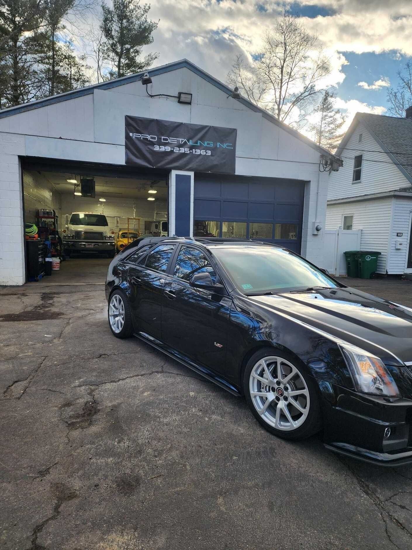 Black Cadillac CTS-V parked in front of a garage, white wheels, open garage bay, sunny outdoor setting.