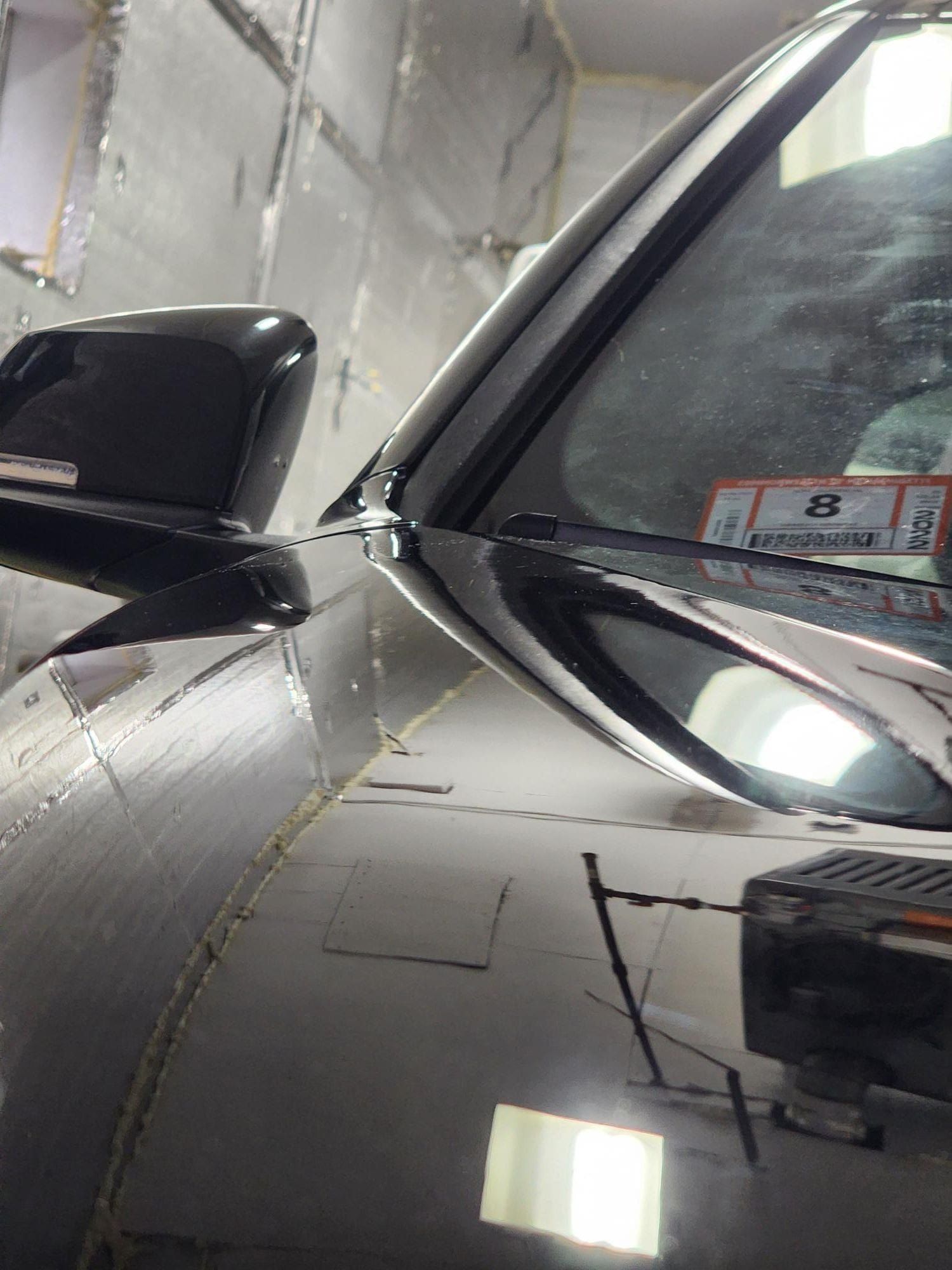 Close-up of a shiny black car's hood, side mirror, and windshield; indoors with reflective walls.