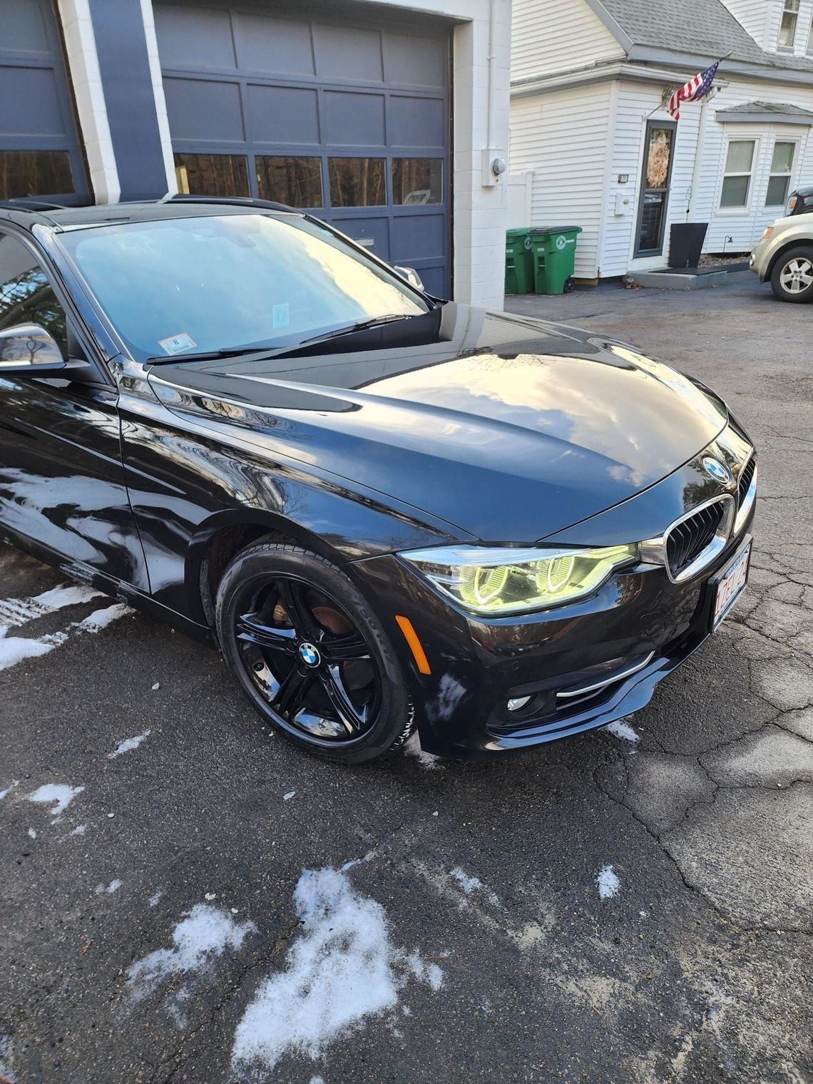 Black BMW parked in front of a garage with yellow tinted headlights, snow on the ground.