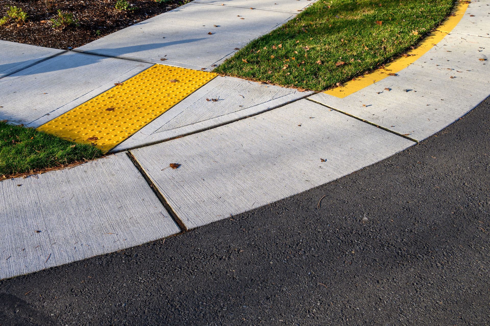 A concrete curb ramp with yellow tactile paving for accessibility, transitioning from a sidewalk to an asphalt road.