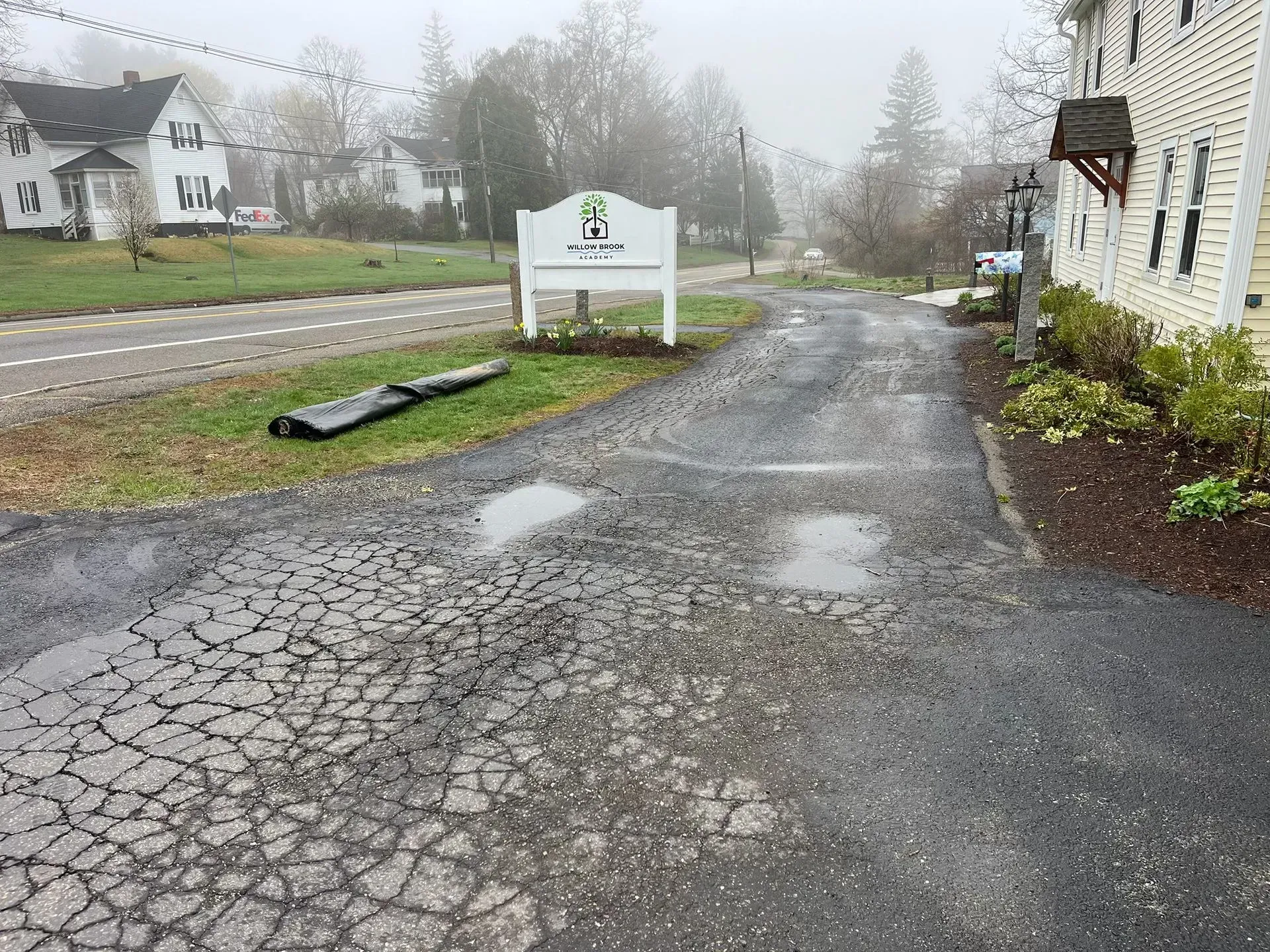 Cracked asphalt driveway leads to a sign in front of a building on a foggy day.