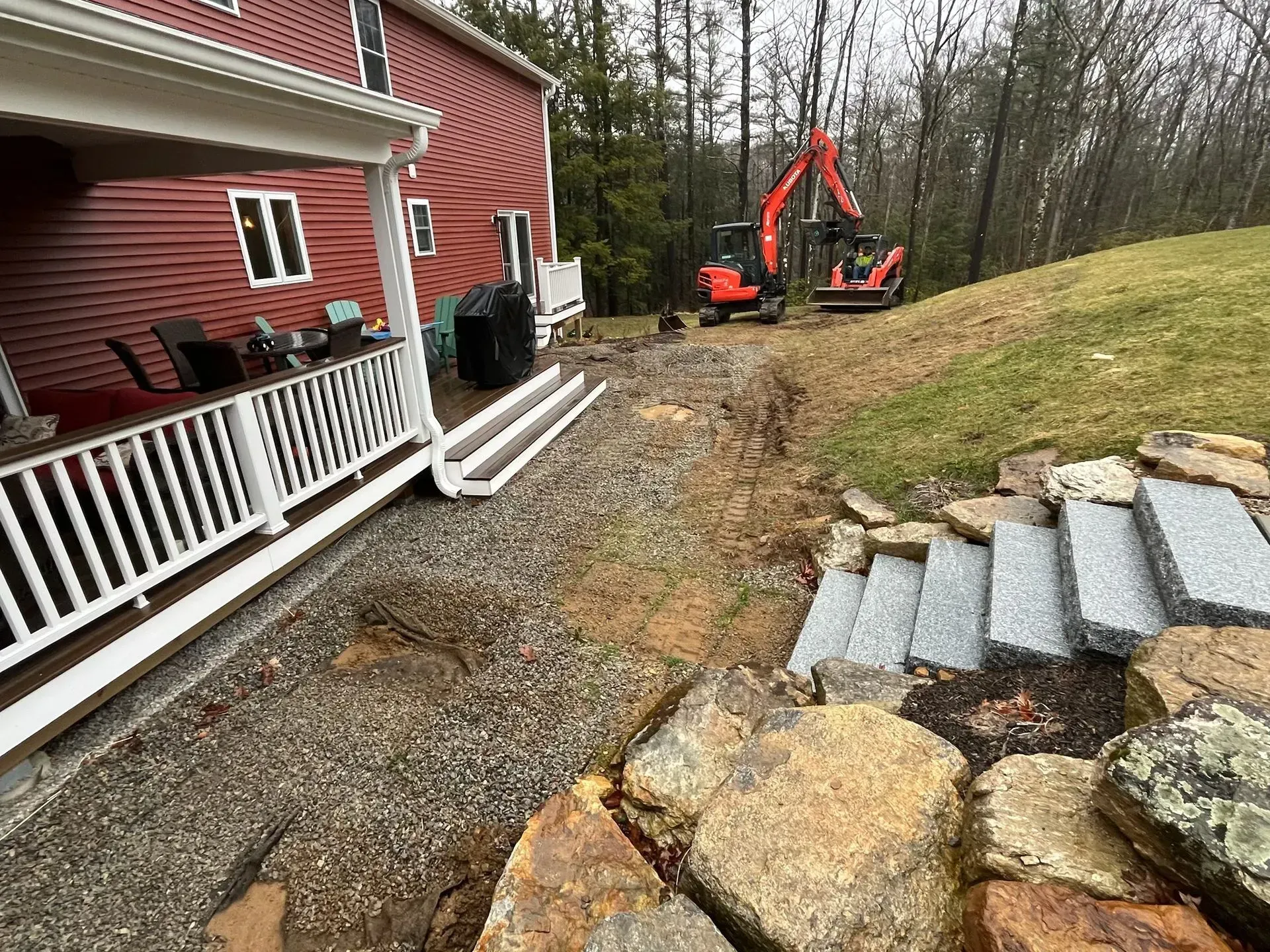 Construction site: Red house with porch, drainage trench, mini excavator, hillside stairs.