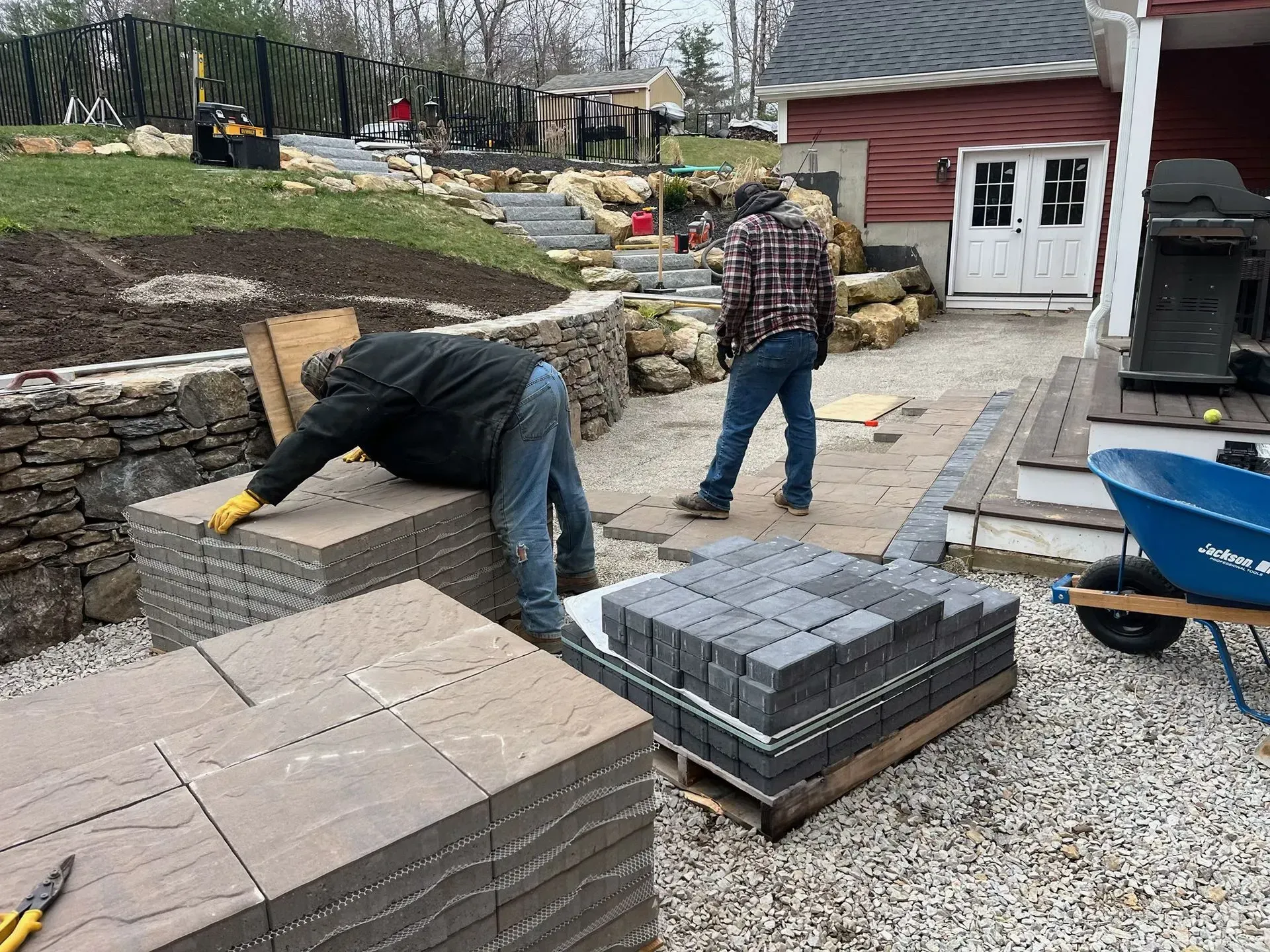 Workers laying pavers outdoors; stacks of stone, wheelbarrow, building construction.