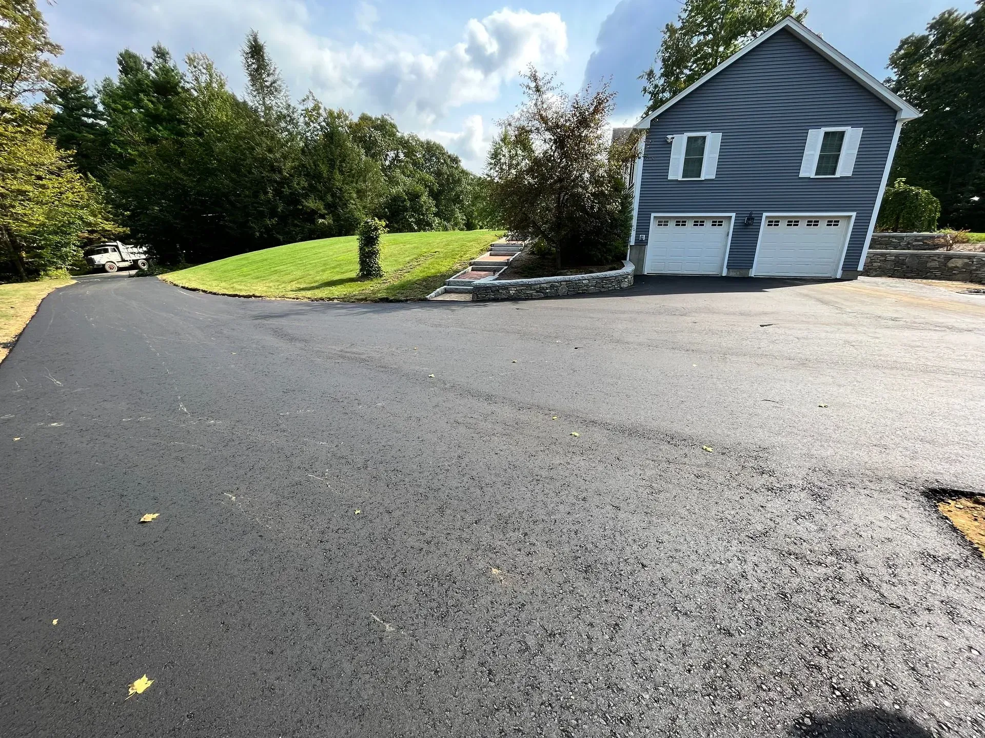 Newly paved black asphalt driveway leading to a blue house with white garage doors; sunny day.
