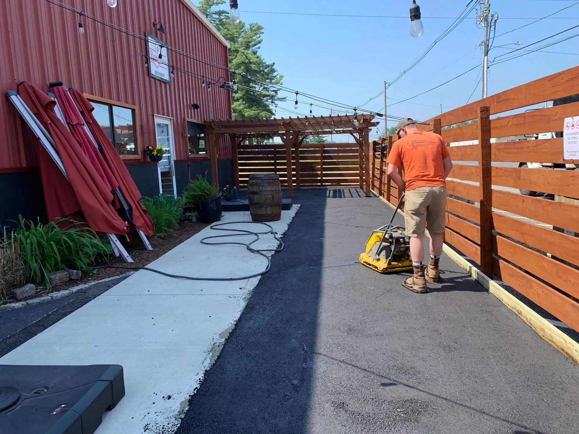 A person compacts asphalt on a patio next to a red building with string lights and a wooden fence.