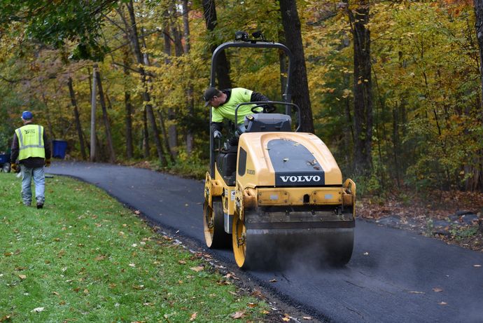 Road crew paving a forest path with a yellow Volvo roller.