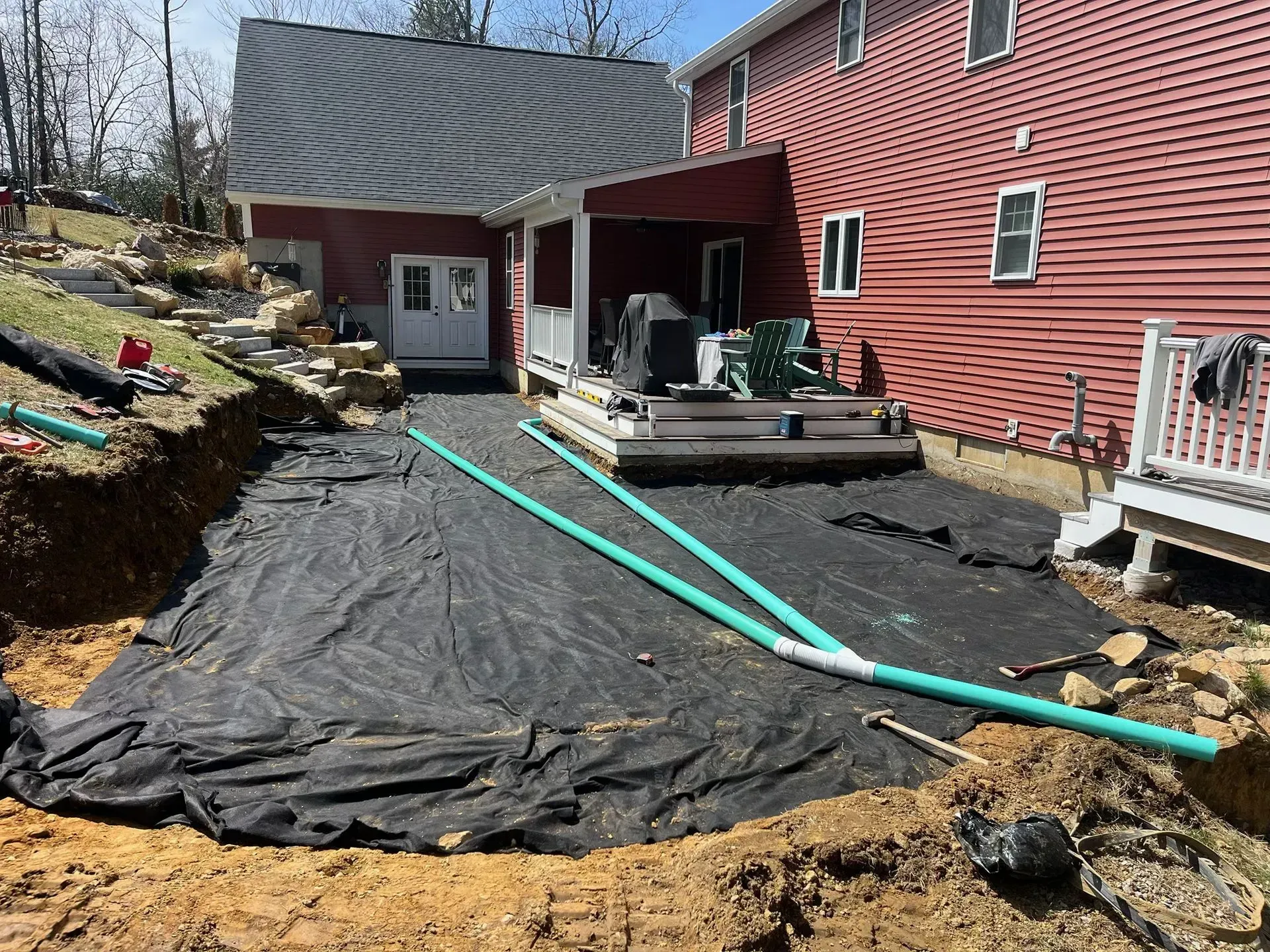 Backyard construction site beside a red house, with black tarp, exposed soil, and turquoise pipes.