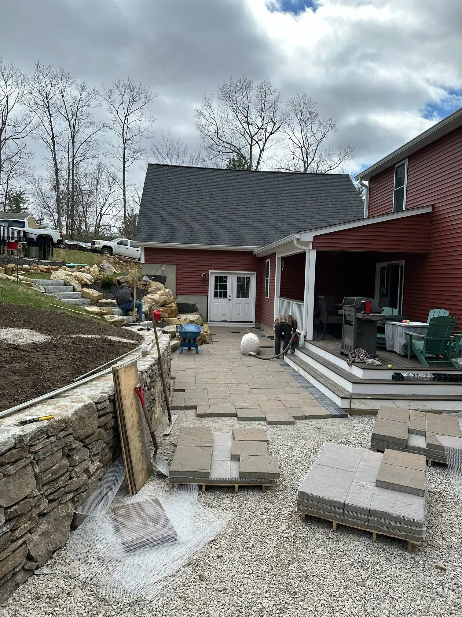 Backyard patio with stone steps, gravel, red house, and stacked pavers under cloudy sky