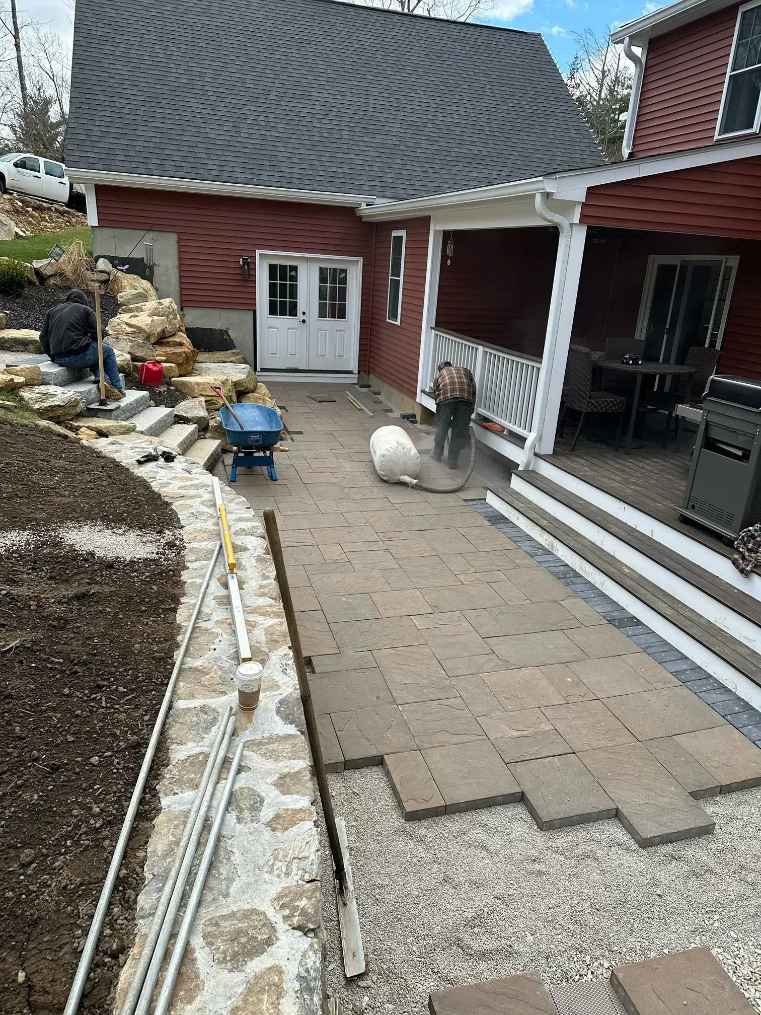 Backyard patio under construction beside a red house, with pavers, gravel, tools, and stone edging