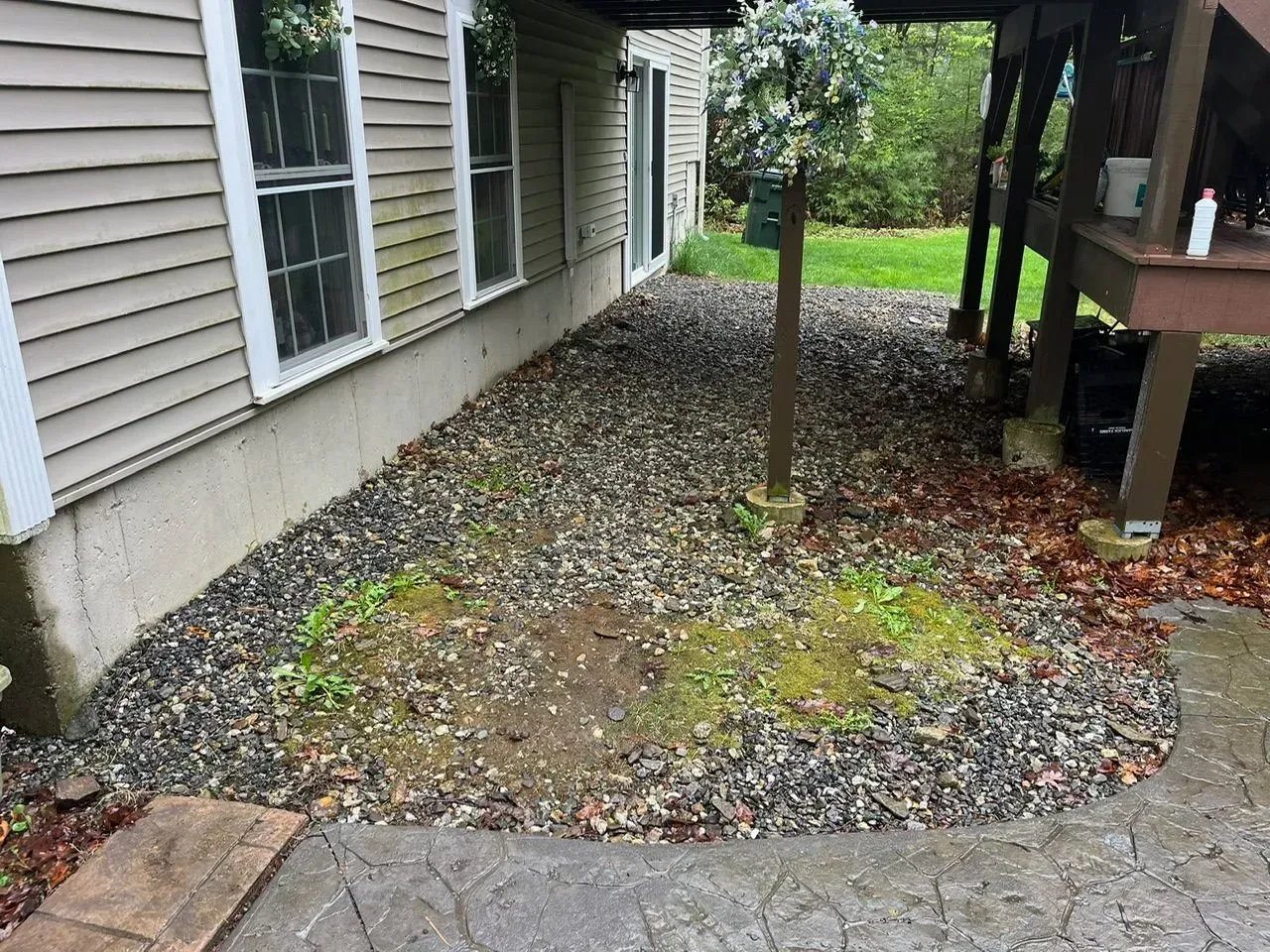 Gravel-covered area between a house and deck, with visible windows. Greenery and a post are also visible.