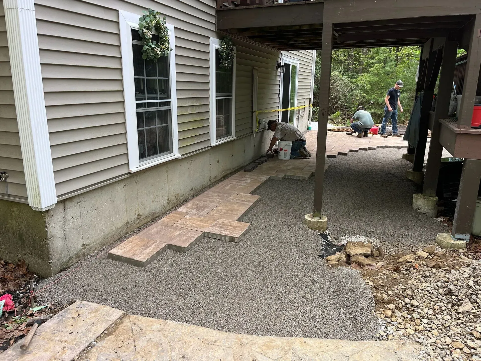 Workers laying stone pavers on a gravel surface next to a house under a deck.