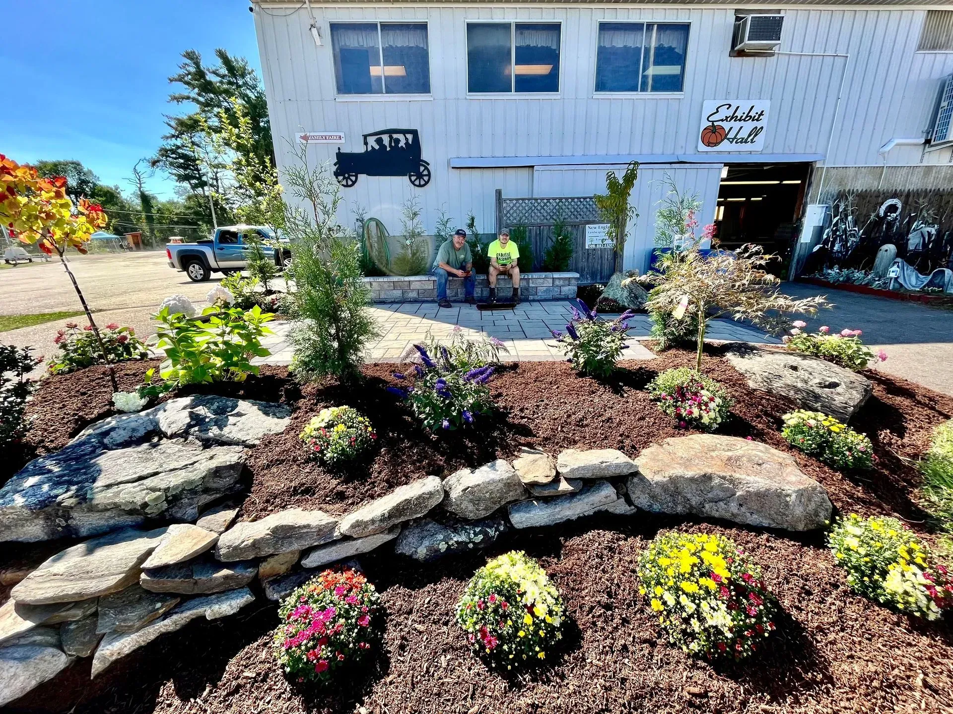Landscaped garden with flowers in front of a white building with a truck logo. Two people are near the entrance.