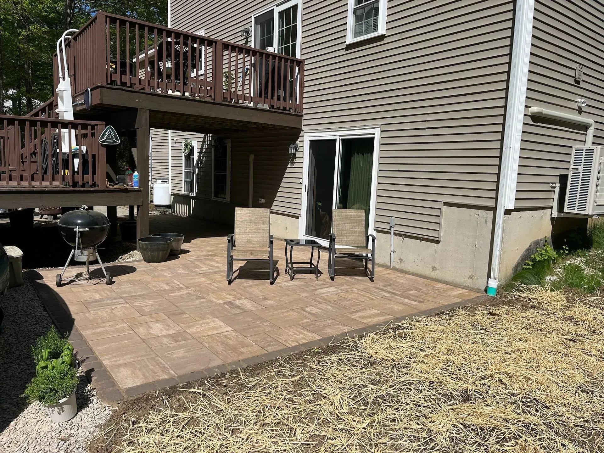 Backyard patio with chairs, table, grill, and deck attached to a house with beige siding.
