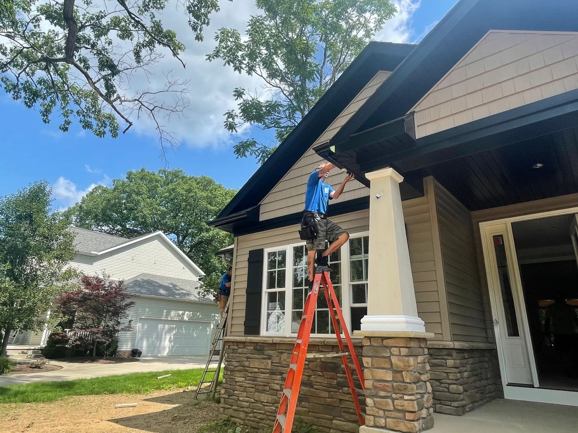 Person on orange ladder trimming tree branches beside a beige house under a blue sky