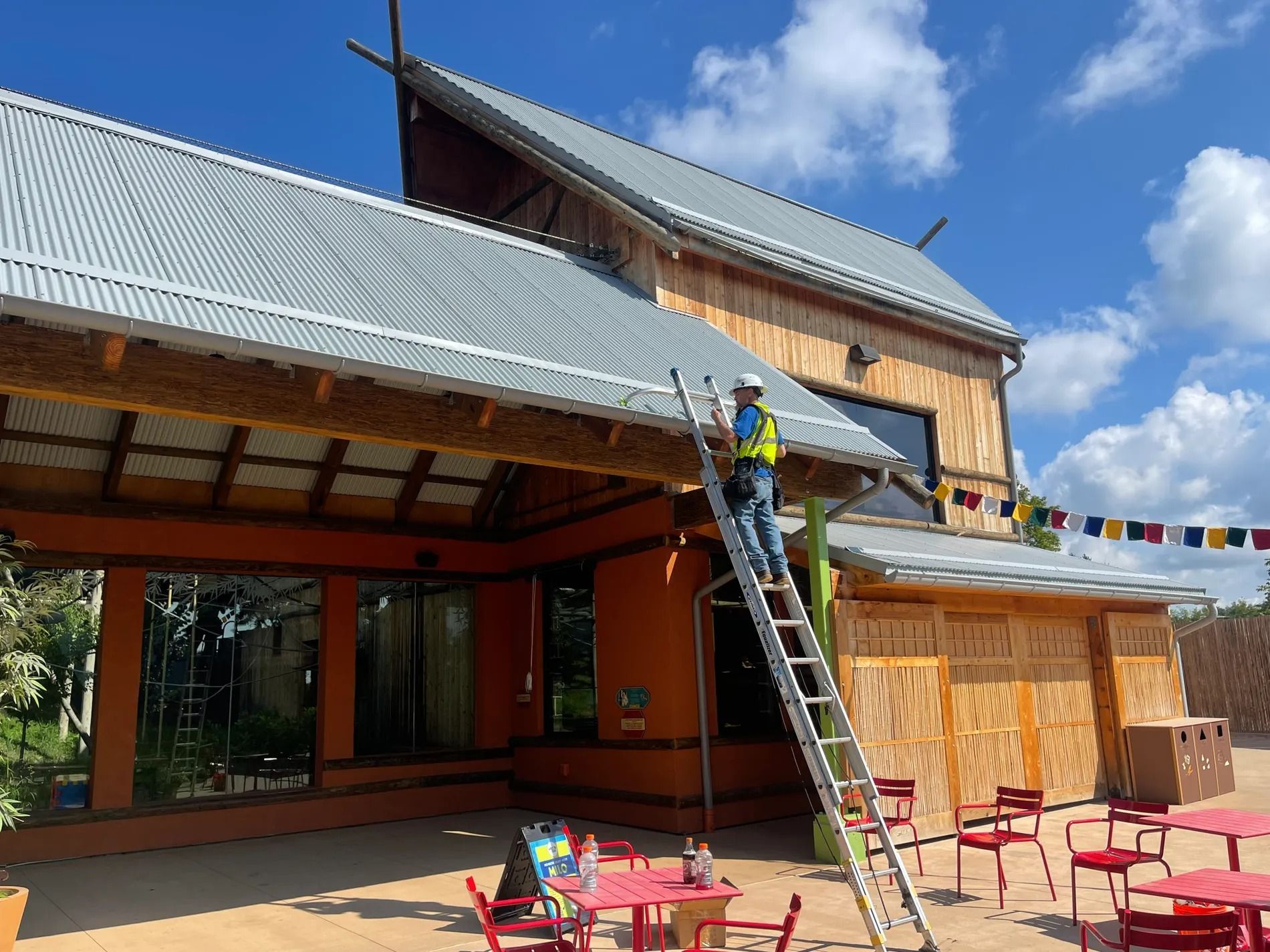 Worker on ladder painting a wooden building with a metal roof, outdoor patio furniture below.