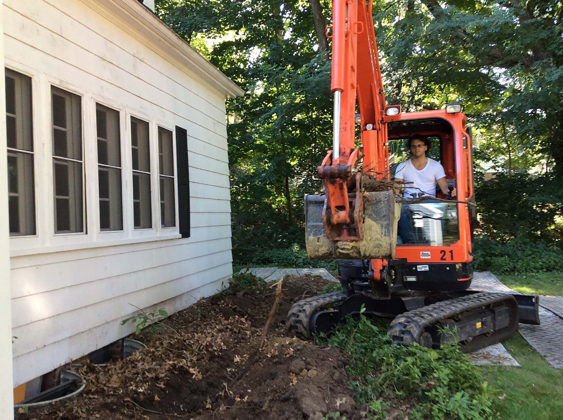 A man is driving an orange excavator in front of a white house.