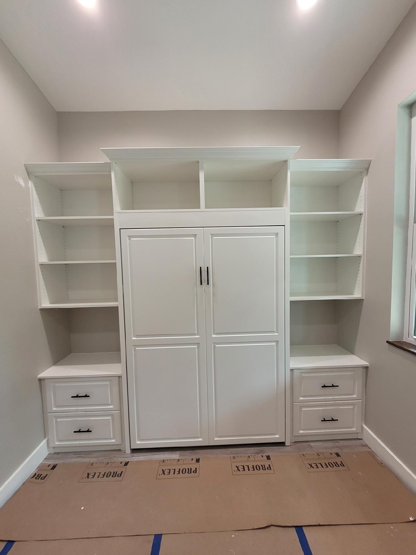 A room with a wall bed , shelves , drawers and a window.