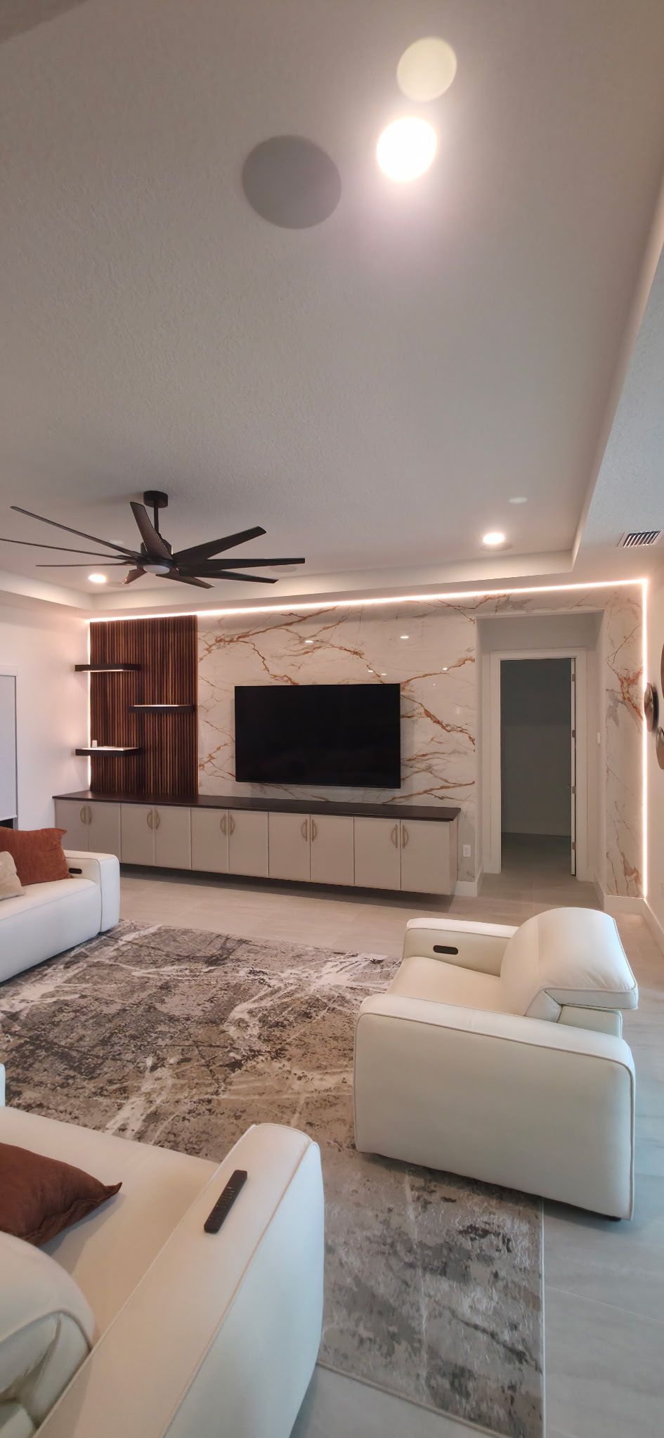 A modern living room with cream furniture, a marble accent wall with a flat-screen TV, a ceiling fan, and tiled floors. 