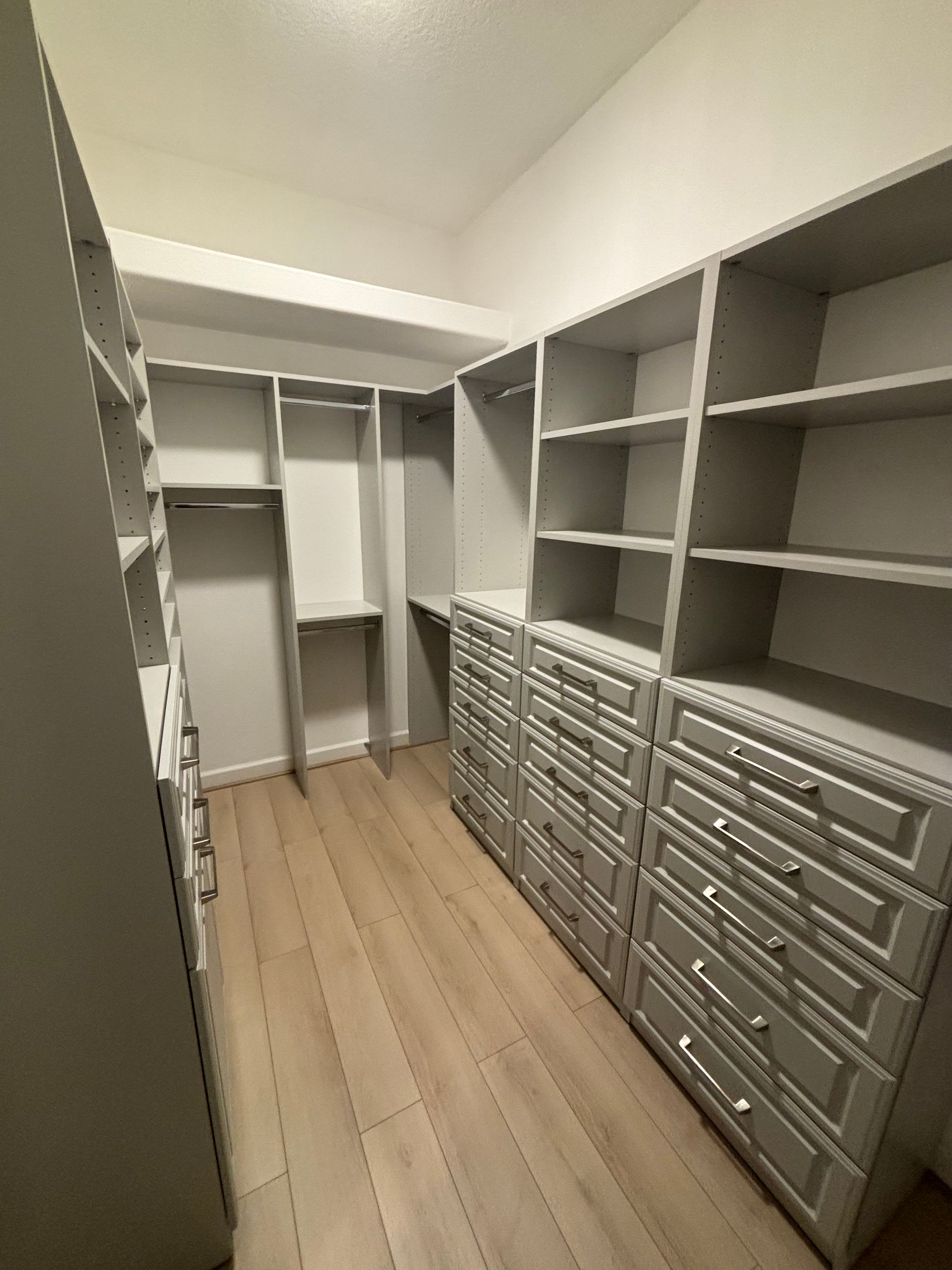 A walk-in closet with light grey custom shelving, multiple drawers, hanging rods, and light wood-tone flooring. 