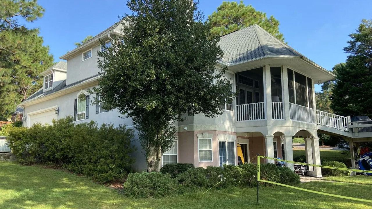 A large white house with a black roof is surrounded by trees and bushes.