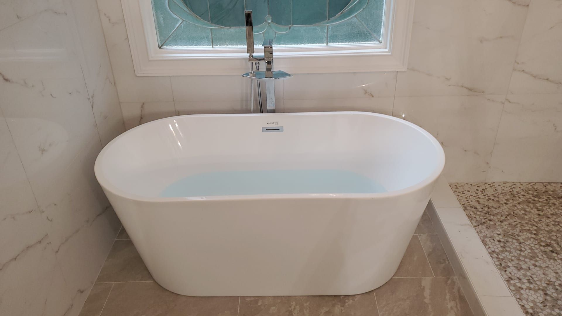 Modern freestanding white bathtub beneath a window in a tiled bathroom