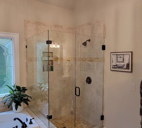 Walk-in shower with glass doors in a beige tiled bathroom, beside a window and potted plant