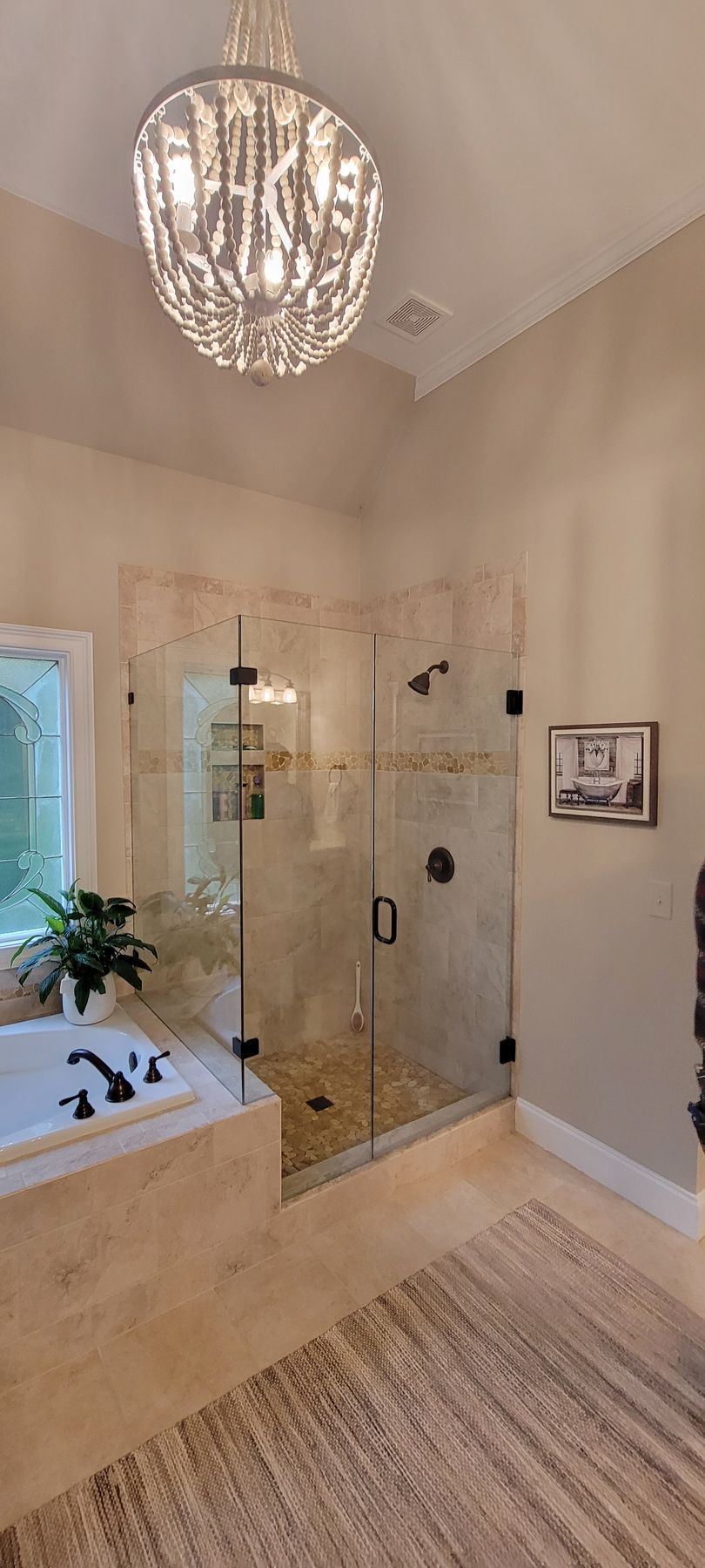 Bright bathroom with a glass shower, soaking tub by a window, chandelier, and woven rug.